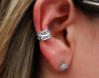 May include: A silver ear cuff with a detailed design. The cuff is worn on the upper part of the ear, just above the lobe.