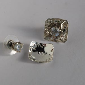 May include: A pair of silver earrings with a textured square design and a clear rhinestone in the center.