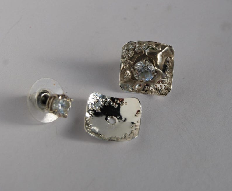 May include: A pair of silver earrings with a textured square design and a clear rhinestone in the center.