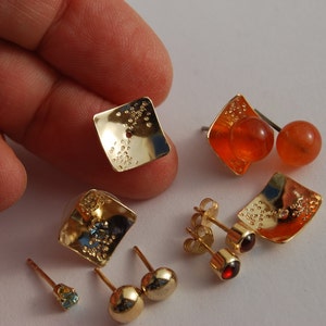 May include: A set of gold earrings with different colored stones. The earrings are square shaped and have a textured surface. One pair has a blue stone, one pair has a red stone, and one pair has an orange stone.
