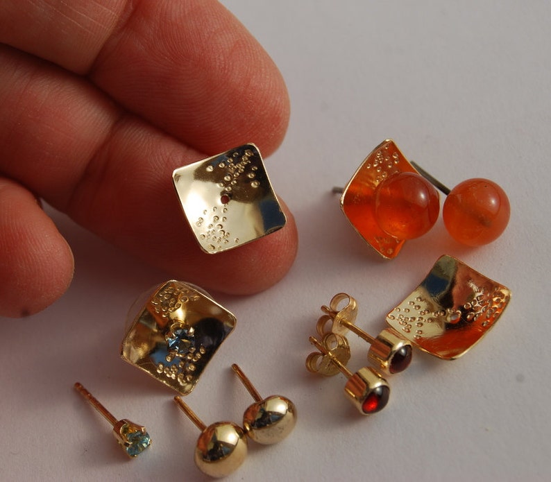 May include: A set of gold earrings with different colored stones. The earrings are square shaped and have a textured surface. One pair has a blue stone, one pair has a red stone, and one pair has an orange stone.