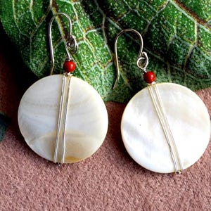 May include: A pair of white shell earrings with silver wire accents and red beads. The earrings are round and have a natural, organic look.