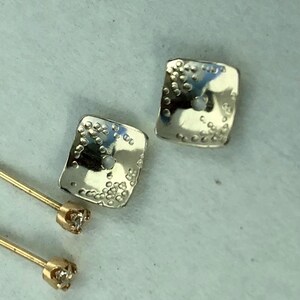 May include: A pair of silver square earrings with a textured surface and a small diamond on each post. The earrings are on a white surface.