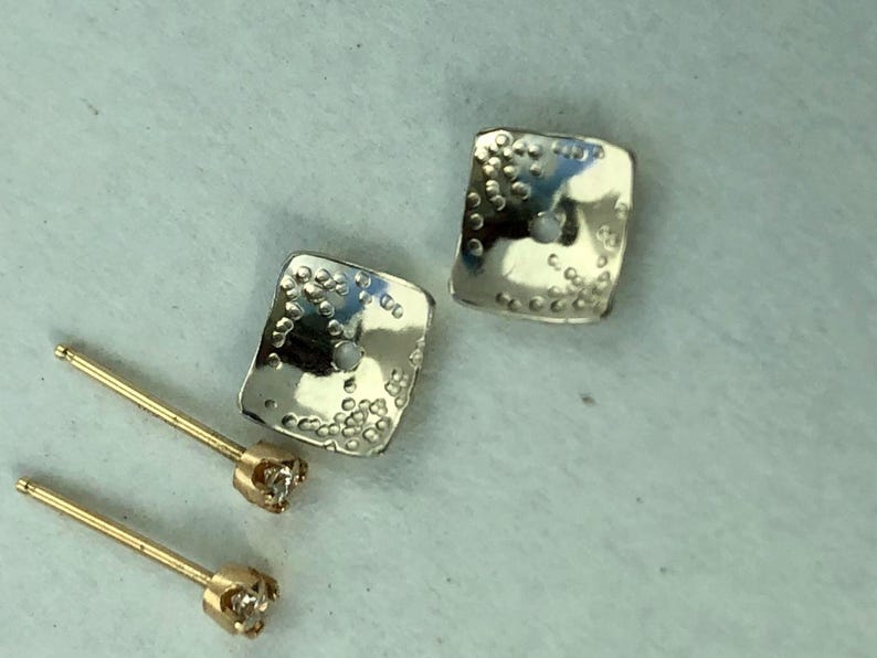 May include: A pair of silver square earrings with a textured surface and a small diamond on each post. The earrings are on a white surface.