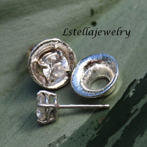 Puede incluir: Un par de pendientes de plata con una piedra preciosa transparente engastada en un engaste de plata texturizado. Los pendientes se muestran sobre un fondo verde.