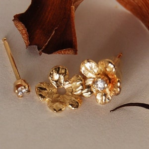 May include: A pair of gold-toned flower stud earrings featuring a textured petal design and a central clear stone. The set includes a separate gold post with a clear stone and a detachable gold flower-shaped jacket component for the earring.