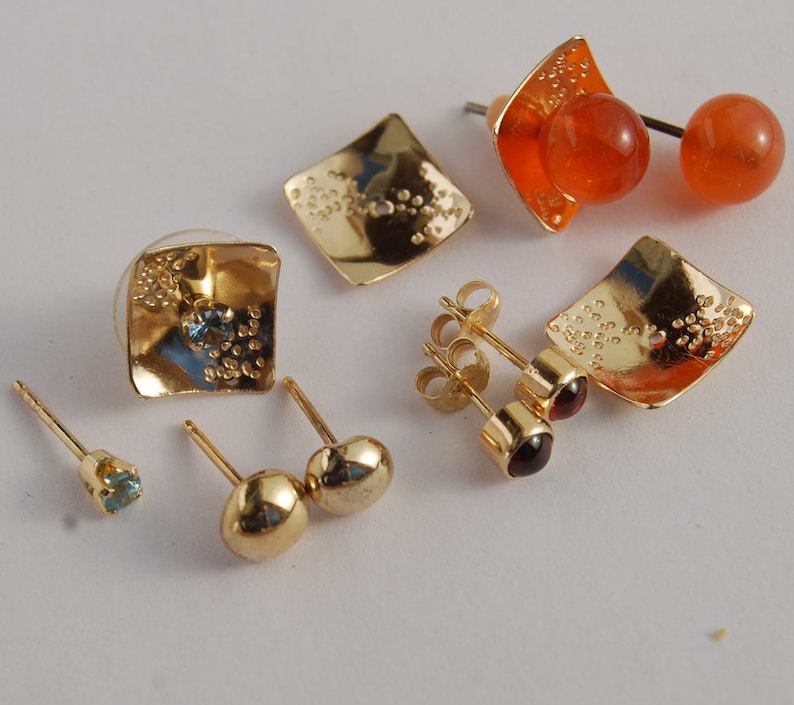 May include: A set of six gold earrings with various gemstones. The earrings feature a variety of styles, including square, round, and teardrop shapes. The gemstones are blue, red, and orange.