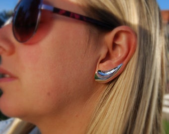 May include: A close-up of a woman's ear with a silver ear cuff. The cuff is shaped like a crescent moon and is worn on the upper part of the ear.