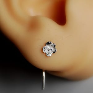 Puede incluir: Un pendiente de flor de plata con un diseño pequeño y delicado. El pendiente se muestra en primer plano, con el pendiente de flor en el centro de la imagen.