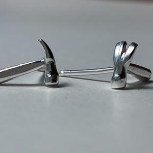 May include: A pair of silver-colored cufflinks in the shape of small hammers. The hammer heads are detailed, and the handles are smooth. The cufflinks are displayed on a light gray surface.