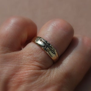 K&ouml;nnte beinhalten: Ein goldener Ring mit einem detaillierten Design auf der Oberseite. Der Ring wird an einem Finger getragen.