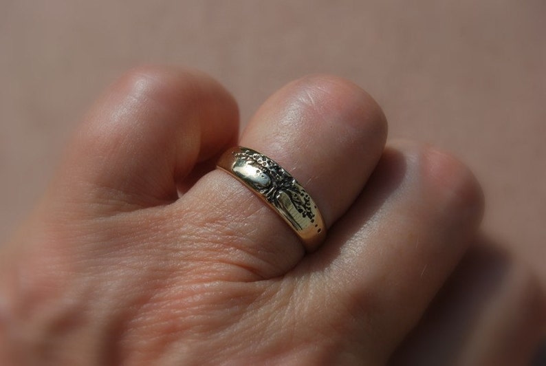K&ouml;nnte beinhalten: Ein goldener Ring mit einem detaillierten Design auf der Oberseite. Der Ring wird an einem Finger getragen.