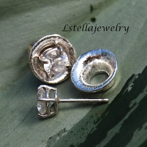 May include: A pair of silver earrings with a clear gemstone set in a textured silver setting. The earrings are shown on a green background.