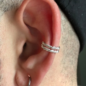 Puede incluir: Un pendiente de aro de plata con un diseño de cuerda trenzada. El aro se lleva en la parte superior de la oreja, por encima del lóbulo.
