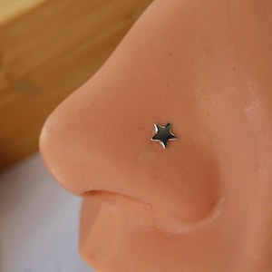 May include: A small, polished silver-toned star-shaped nose stud. The jewelry features a five-pointed star design with a smooth, reflective metallic surface, positioned securely on the side of a nose model for display purposes.