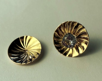 May include: Two circular gold-toned earring jackets with a radial, sunburst-like pleated texture. One jacket features a round, faceted, light-reflecting gemstone set in the center, while the other is a plain, empty disc with a central hole.