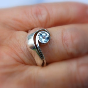 Puede incluir: Un anillo de plata con una piedra preciosa azul engastada en un diseño en forma de ola.