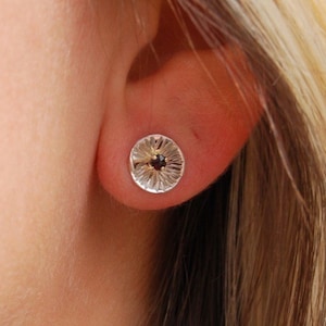 May include: A pair of silver stud earrings with a textured, circular design. Each earring features a small, dark red gemstone in the centre.