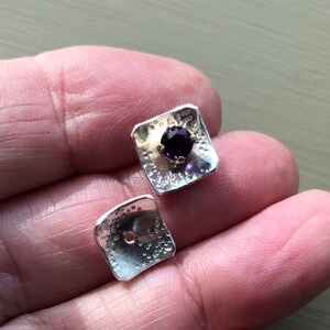 May include: A pair of silver square earrings with a textured surface. One earring features a purple gemstone set in a gold setting.