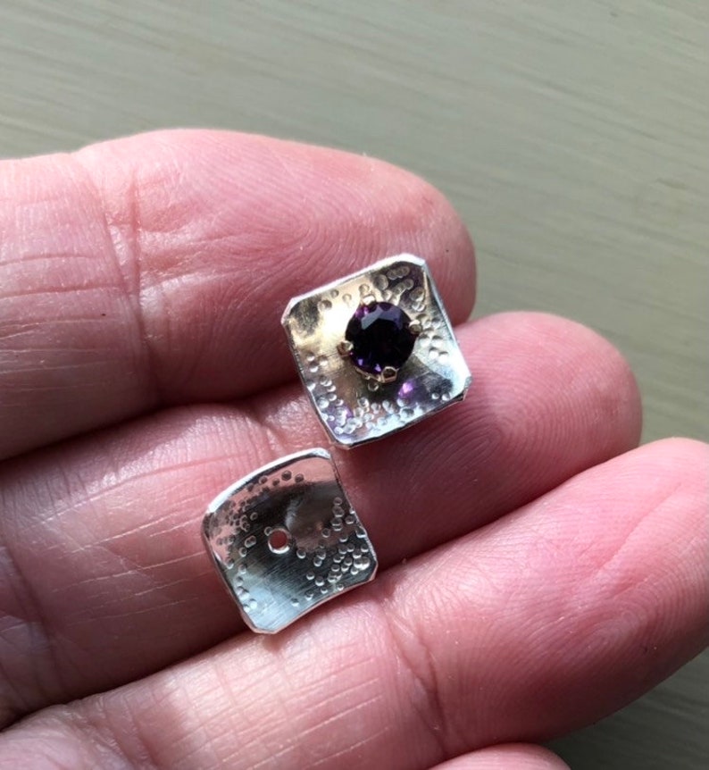 May include: A pair of silver square earrings with a textured surface. One earring features a purple gemstone set in a gold setting.