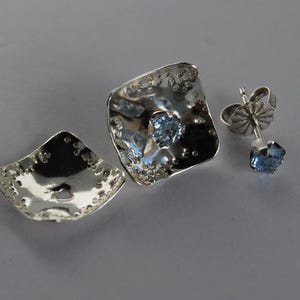 May include: A pair of silver earrings with a blue gemstone in the center of each earring. The earrings are square shaped and have a textured surface.