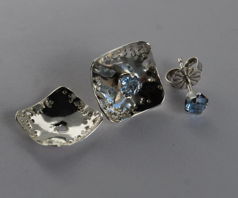 May include: A pair of silver earrings with a blue gemstone in the center of each earring. The earrings are square shaped and have a textured surface.