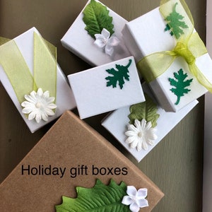 K&ouml;nnte beinhalten: Sortiment kleiner wei&szlig;er Geschenkboxen mit gr&uuml;nen Blatt- und wei&szlig;en Blumenverzierungen. Die Boxen sind auf einer braunen Papieroberfl&auml;che mit dem Text "Holiday gift boxes" angeordnet.