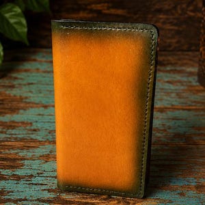 May include: A vertical rectangular leather wallet featuring a gradient color scheme that transitions from a bright amber orange center to deep forest green edges, finished with visible dark green stitching along the perimeter of the folded edges.