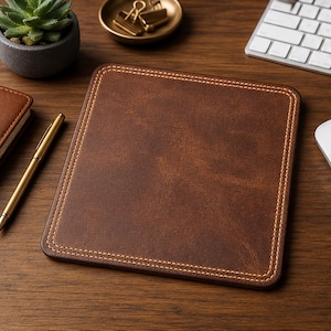 May include: A square brown leather mouse pad featuring rounded corners and decorative light-colored stitching along the entire perimeter. The surface has a textured, matte finish, providing a clean and uniform appearance for a desk workspace.