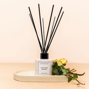 May include: A white ceramic reed diffuser with black reeds, labelled "Leather Books" on a matching tray. The diffuser is surrounded by greenery and spices. The product is from Simon James Home, handmade in Kent.