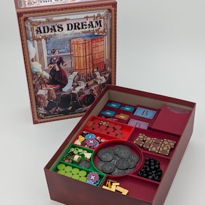 May include: A board game box set titled "Ada's Dream" with a maroon box and a detailed illustration. The open box reveals game pieces in various colors, including green, red, and black, with coins and tokens.