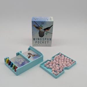 May include: A Wingspan Pocket board game box featuring bird illustrations, a light blue plastic card holder with small coloured wooden tokens, and a light blue tray filled with numerous small, speckled light pink egg-shaped game pieces.
