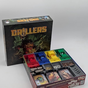 May include: A board game box titled Drillers with a dark, illustrated cover featuring a mechanical character. The open box base contains organised plastic inserts in red, blue, yellow, and green, holding various game cards, tokens, and pieces.
