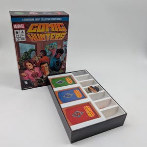 May include: The Marvel Comic Hunters card game box and open tray. The box cover features the title, player count, age, and time icons, with an illustration of people reading comics. The tray contains three stacks of cards and a circular token.