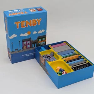 May include: A blue board game box titled TENBY with a cartoon town illustration, next to an open box base containing yellow plastic trays filled with stacks of colourful rectangular cards and small circular tokens in various bright colours.