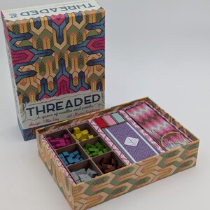May include: A board game titled Threaded featuring a box with a colourful geometric pattern. The open box contains six compartments filled with small wooden cubes in pink, yellow, blue, green, brown, and grey, plus decks of cards and game tiles.
