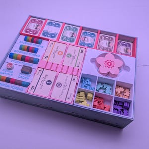Può includere: Un organizer per giochi da tavolo con vari componenti. La scatola contiene carte, gettoni e pedine di diversi colori. Le carte sono in divisori rosa. I gettoni sono etichettati "Revenue", "Offering", "Stock" e "Station". Un fiore di legno è al centro.