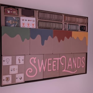 Può includere: Un organizer per giochi da tavolo con scomparti per carte, pedine e altri componenti. L'organizer presenta un design a blocchi di colore con la scritta "SWEETLANDS" in rosa. Gli scomparti sono dipinti in verde, blu, rosso e giallo.