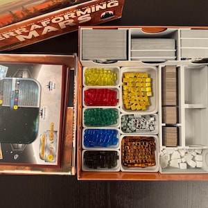 May include: A Terraforming Mars board game box with an open organizer tray containing stacks of cards, white plastic bins filled with translucent yellow, red, green, blue, and black cubes, metallic gold and copper cubes, and silver tokens.