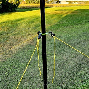 May include: A black pole with yellow ropes extending outwards, set in a grassy field. The pole has a green connector with black clips. The text "DX COMMANDER" is visible on the pole.