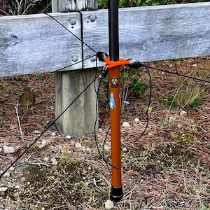May include: An orange and black antenna mast with an orange support structure. The mast is positioned near a wooden fence and is surrounded by natural foliage and rocks. The mast has a black cable attached to it.