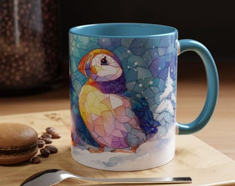 Könnte beinhalten: Eine Keramiktasse mit blauem Innenraum und Griff, die eine farbenfrohe Illustration eines Papageientauchers im Glasmalerei-Stil zeigt. Das Design umfasst eine Winterszene mit Schnee und einem Baum. Ein Löffel und ein Gebäck befinden sich im Vordergrund.