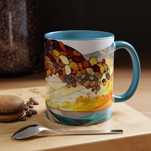 Puede incluir: Una taza de cerámica azul y blanca con un diseño de vitral de una cordillera. La taza tiene un borde y un asa azules.