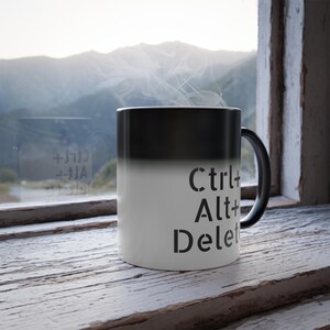 May include: A black and white colour-changing mug with the text "Ctrl+Alt+Delete" printed on the white side. The mug is sitting on a wooden surface in front of a window.