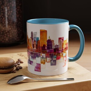 Peut inclure: Une tasse en céramique blanche avec un bord et une anse bleus. La tasse présente un motif de ville aquarelle coloré avec une variété de bâtiments dans des tons d'orange, de violet et de rose.