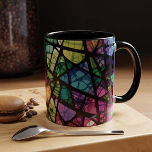 May include: A black coffee mug with a colourful stained glass design. The design features a variety of colours, including blue, green, purple, and yellow.
