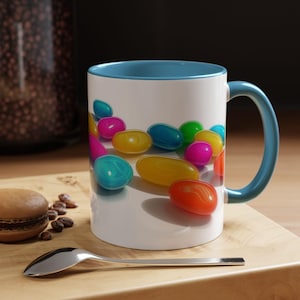 May include: A white ceramic mug with a blue rim and handle. The mug features a colorful design of jelly beans scattered on a white background.
