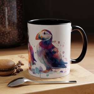 Pode incluir: Caneca de cerâmica branca com interior e alça pretos, com uma ilustração em aquarela de um papagaio-do-mar. A ave tem um bico e plumagem coloridos em tons de roxo, azul e laranja. A caneca está sobre uma superfície de madeira.