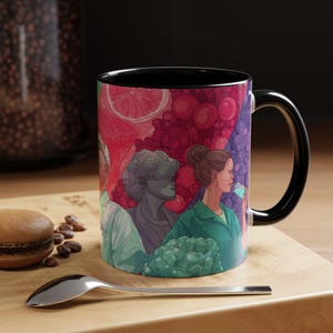 May include: A black-handled coffee mug with a vibrant illustration of figures and fruit. The mug's design features a woman in a green shirt, a statue, and a person in a white coat. The background includes red and purple fruit. The mug is on a wooden surface.