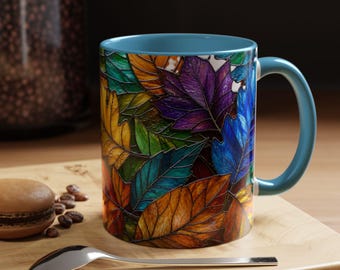 Könnte beinhalten: Ein Keramikbecher mit hellblauem Innenraum und Henkel, der ein lebhaftes Buntglas-Muster aus überlappenden Herbstblättern in Violett, Blau, Gelb, Orange und Grün zeigt, mit einer glänzenden Oberfläche und einem geschwungenen Henkel.
